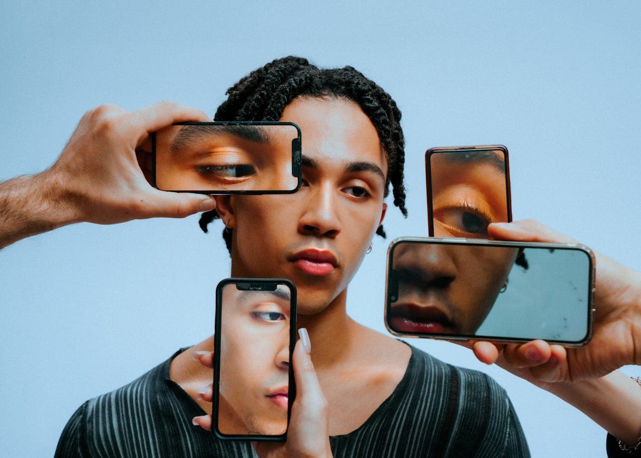 A young man surrounded by hands holding phones that reflect pieces of his face